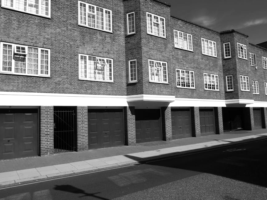 Black and white photograph of a modern three-story residential building with multiple rectangular windows, some with visible window frames and open panes, and brick exterior walls. The ground level features several closed garage doors made of metal, with some sections showing smaller pedestrian gates or entry points. A sidewalk runs in front of the building along a paved street, with shadows cast onto the pavement, indicating sunlight. Part of the moving process may involve loading or unloading furniture or boxes, although no activity or people are visible in the image. The setting suggests an urban residential area prepared for home relocation or furniture transport, with the building design supporting professional removals services such as those offered by Man and a Van Finsbury Park.