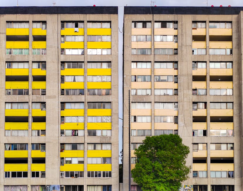 Front view of a multi-storey residential building with a concrete facade, featuring a repetitive pattern of windows and balconies. The balconies on the left side are painted with bright yellow panels, creating a stark contrast against the neutral grey concrete, while the right side shows balconies with beige and cream-colored coverings. Some windows are open or have curtains visible, and a few contain laundry or personal items. In the foreground, a lush green tree partially obscures the lower part of the building. The scene appears to be captured in daylight under overcast conditions, with natural light evenly illuminating the building. This image may relate to property or housing in an urban environment, relevant to house removals or relocation services provided by Man and a Van Finsbury Park, as featured on their website page about removals for homes on Stroud Green Road.