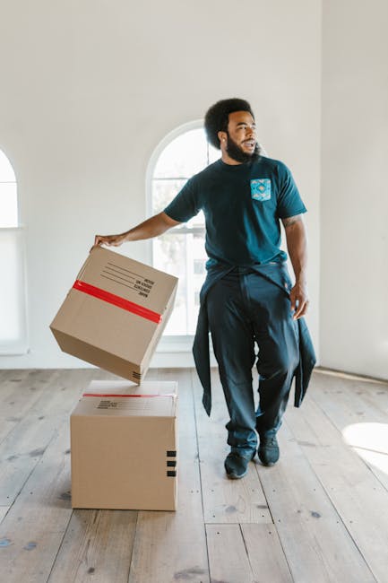 A male mover with dark curly hair and a beard stands inside an uncluttered, well-lit room with white walls and a large arched window, holding a cardboard box wrapped with red packing tape in his right