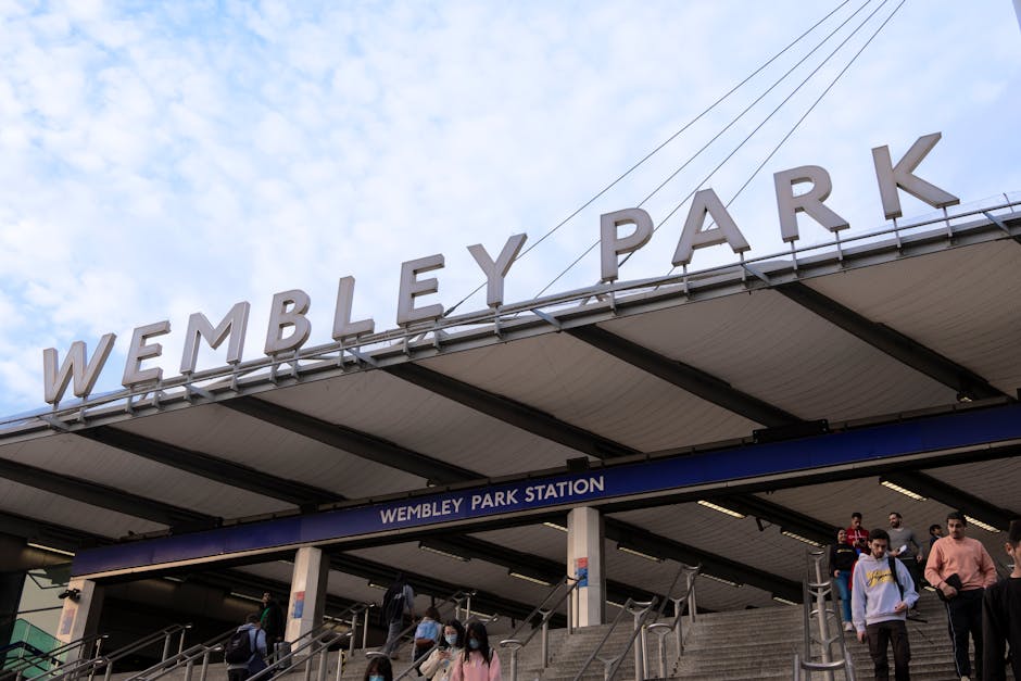The exterior of Wembley Park Station entrance with a large sign displaying the station's name in bold letters against a cloudy sky. The station features a covered walkway with dark metal supports and a roof. Below the sign, there are multiple staircases leading down to the station platform, with a group of people ascending and descending. Some individuals are wearing face masks, carrying backpacks or bags, and dressed in casual clothing. The image captures the urban environment of a busy travel hub associated with house relocations and moving logistics, emphasizing transportation services near Finsbury Park and potential furniture transport activities involved in home removals or packing and moving tasks managed by Man and a Van Finsbury Park.
