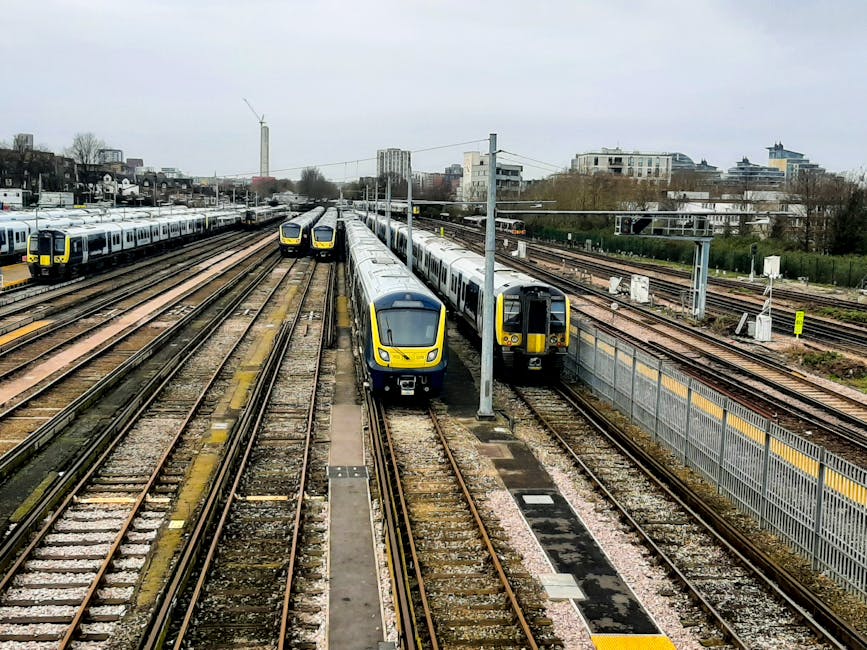 A view of multiple modern passenger trains parked on parallel railway tracks at a station near Finsbury Park, with two trains positioned side by side in the foreground. One train has a sleek, yellow and white front, while another adjacent train is dark grey with yellow accents. The tracks are made of steel rails with wooden sleepers and are surrounded by gravel. Alongside the tracks, there is a tall metal fence with yellow and grey barriers, and overhead electric power lines supported by metal poles run above the trains. In the background, there are urban residential and commercial buildings, some high-rise, under an overcast sky. The scene depicts the infrastructure typically involved in train and transportation logistics, relevant to house removals or moving services by companies like Man and a Van Finsbury Park, who coordinate the transportation and logistical aspects of home relocation and furniture transport.